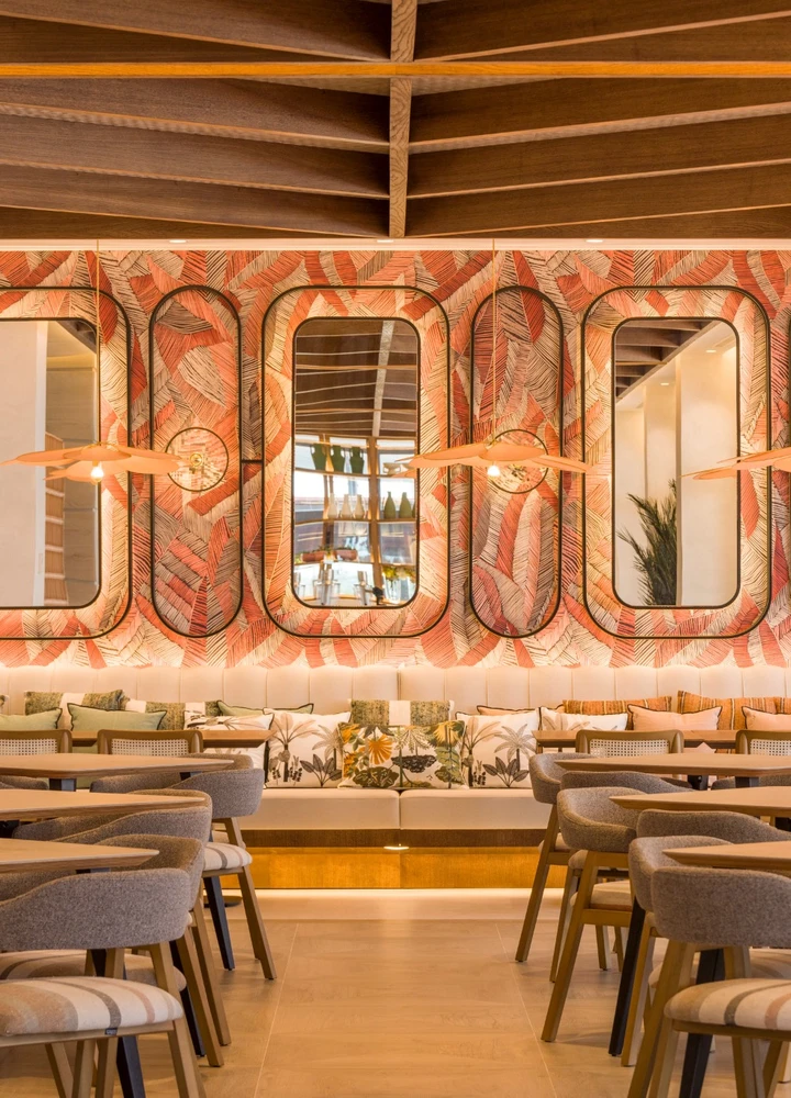 Interior del restaurante del hotel con mesas de madera y sillas tapizadas, banco corrido acolchado y una pared decorada con papel floral en tonos rojos y neutros. Sobre la pared, varios espejos verticales con marcos redondeados reflejan una barra con botellas. El techo presenta un diseño de madera con vigas y la iluminación cálida incluye lámparas colgantes y luz indirecta.