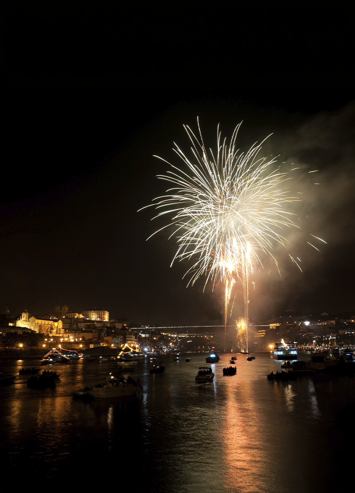 Fuegos artificiales dorados explotando en el cielo nocturno sobre un puerto, con varias embarcaciones dispersas en el agua que refleja los destellos de luz; al fondo, una ciudad iluminada con edificios históricos en una colina y un puente que cruza de lado a lado, mientras una nube de humo se expande alrededor de las explosiones.