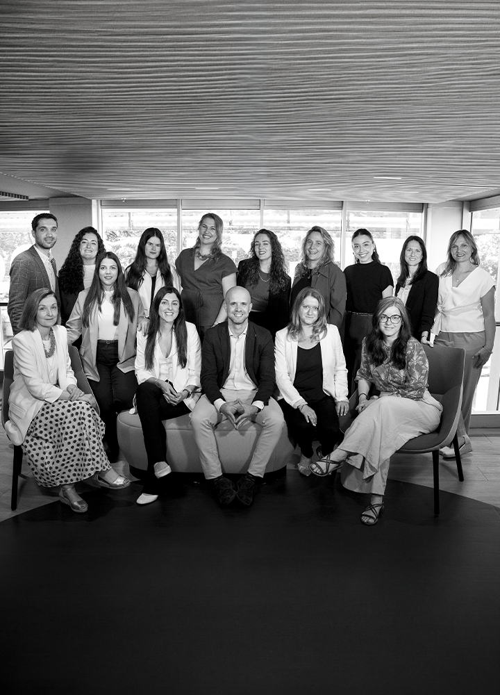 Fotografía de grupo en blanco y negro con quince personas posando en el interior de una sala moderna. Algunas están sentadas y otras de pie alrededor de un sofá circular. Al fondo hay grandes ventanales que dejan entrar luz natural y un techo con revestimiento de madera. La composición está centrada en el grupo, con mobiliario contemporáneo y un ambiente corporativo o profesional.