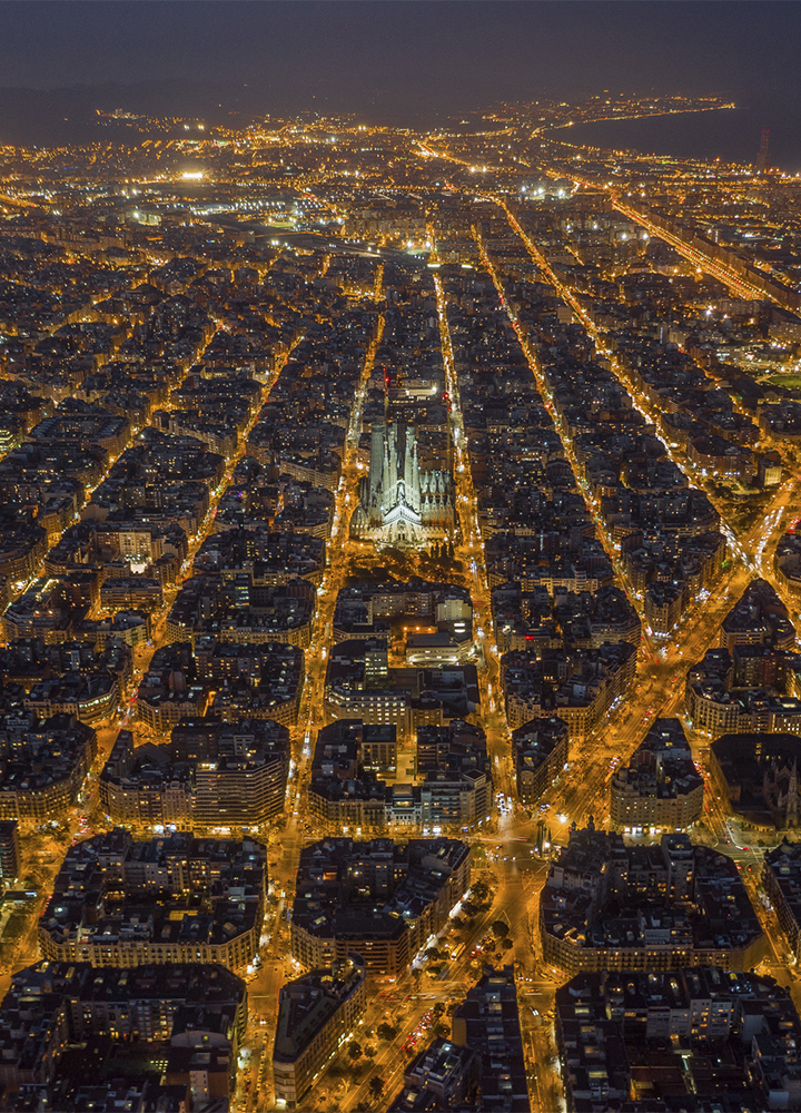 Vista aérea nocturna de una ciudad con cuadrícula muy regular y manzanas octogonales iluminadas por luces cálidas; en el centro destaca una gran basílica de varias torres claramente iluminada y, hacia la derecha, un rascacielos moderno con iluminación de color rompe la uniformidad, mientras la trama urbana se extiende hasta el horizonte donde se intuyen colinas y una masa de agua en la oscuridad.