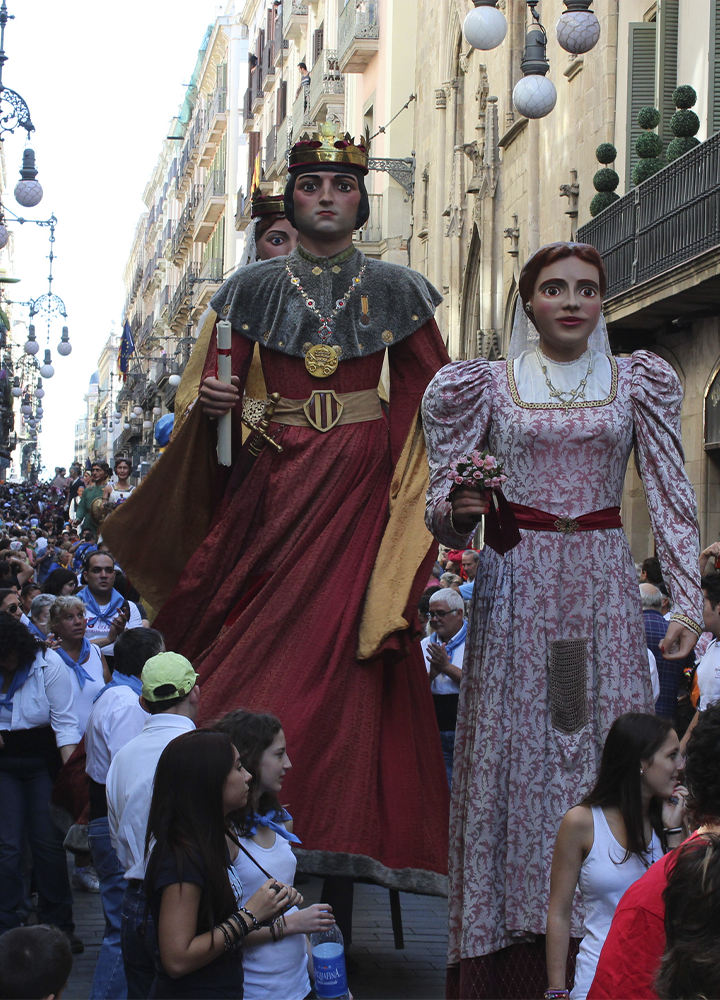 Desfile multitudinario por una calle estrecha flanqueada por edificios con balcones y farolas, donde avanzan dos enormes figuras festivas de tamaño gigante vestidas con ropa histórica: una con túnica roja, capa y corona que sostiene un cetro, y otra con vestido claro estampado y ramo en la mano; ambas están rodeadas de una gran cantidad de personas que caminan y observan desde la calle y los laterales, creando un ambiente de celebración urbana a plena luz del día.