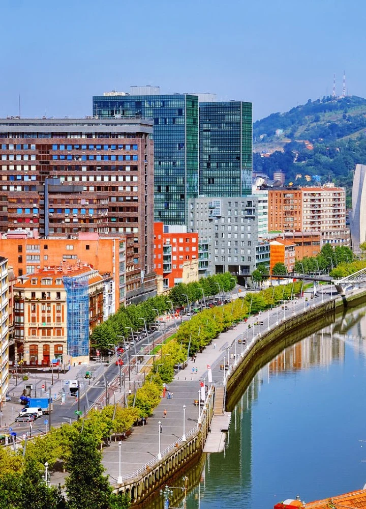 Vista urbana junto a un río de aguas tranquilas que curva hacia la derecha. En primer plano, un paseo peatonal amplio con baldosas claras, árboles alineados y farolas blancas; aparecen varias personas caminando y zonas de descanso. A la izquierda del paseo se alzan edificios de distintos estilos: uno bajo de fachada clásica en tonos crema y teja naranja, otro alto con fachada marrón y numerosas ventanas, y varios bloques modernos con vidrio verde y gris. Al fondo, una colina cubierta de vegetación con antenas en la cima. Cielo despejado y luminoso, con luz diurna que resalta los colores de los edificios y la vegetación.