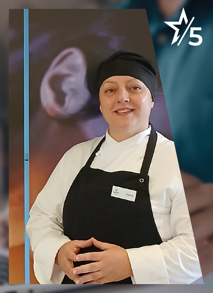 Persona vestida con uniforme de cocina compuesto por chaqueta blanca, delantal negro y gorro oscuro posa de pie con las manos entrelazadas a la altura del abdomen, sobre un fondo desenfocado donde destaca una imagen ampliada de una oreja; en la esquina superior derecha aparece un icono de estrella acompañado del número “5”, y en el pecho se aprecia una pequeña placa identificativa.