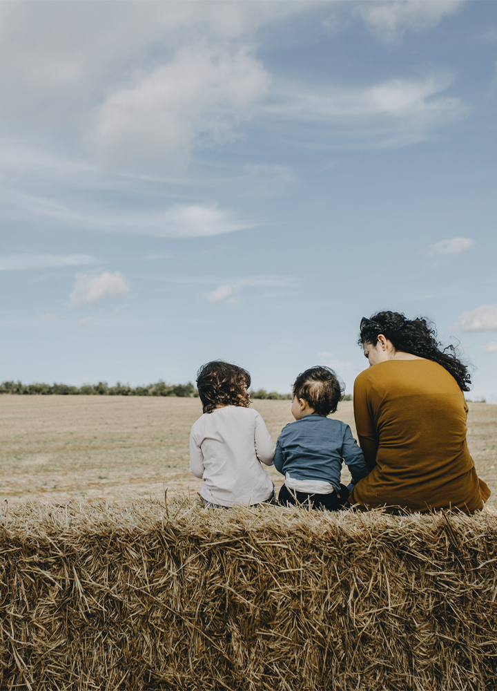 Escena al aire libre en un campo abierto bajo un cielo amplio con nubes dispersas, donde tres personas aparecen sentadas de espaldas sobre un gran fardo de paja en primer plano; las dos figuras más pequeñas se sitúan en el centro con ropa informal de tonos claros y azul, mientras la figura situada a la derecha viste una prenda de color marrón y tiene el cabello recogido, todas apoyadas mirando hacia la extensión del terreno; frente a ellas se extiende una llanura de tonos dorados y tierra seca con una línea de árboles en el horizonte, y la luz suave resalta las texturas de la paja y el paisaje, generando una composición tranquila y equilibrada sin presencia de texto ni elementos adicionales.