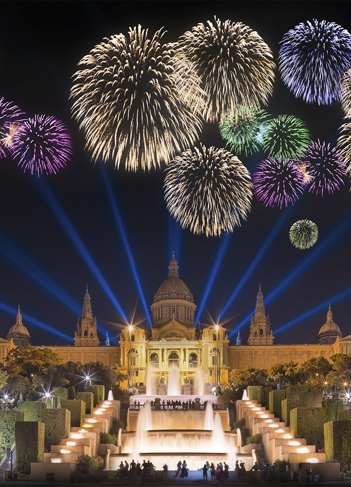 Espectáculo nocturno de fuegos artificiales de varios colores —dorados, morados, verdes y azules— que explotan en el cielo sobre un gran edificio histórico iluminado con luz cálida, con una cúpula central y dos torres laterales; potentes haces de luz azul se proyectan hacia arriba desde el suelo, mientras en primer plano se alinean fuentes escalonadas y zonas ajardinadas con setos geométricos, y pequeñas siluetas de personas observan el evento desde la parte inferior.