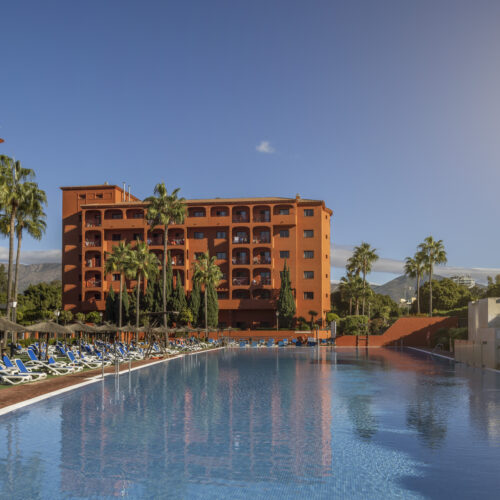 Complejo hotelero con edificios terracota alrededor de una gran piscina, rodeada de hamacas y palmeras bajo cielo despejado.