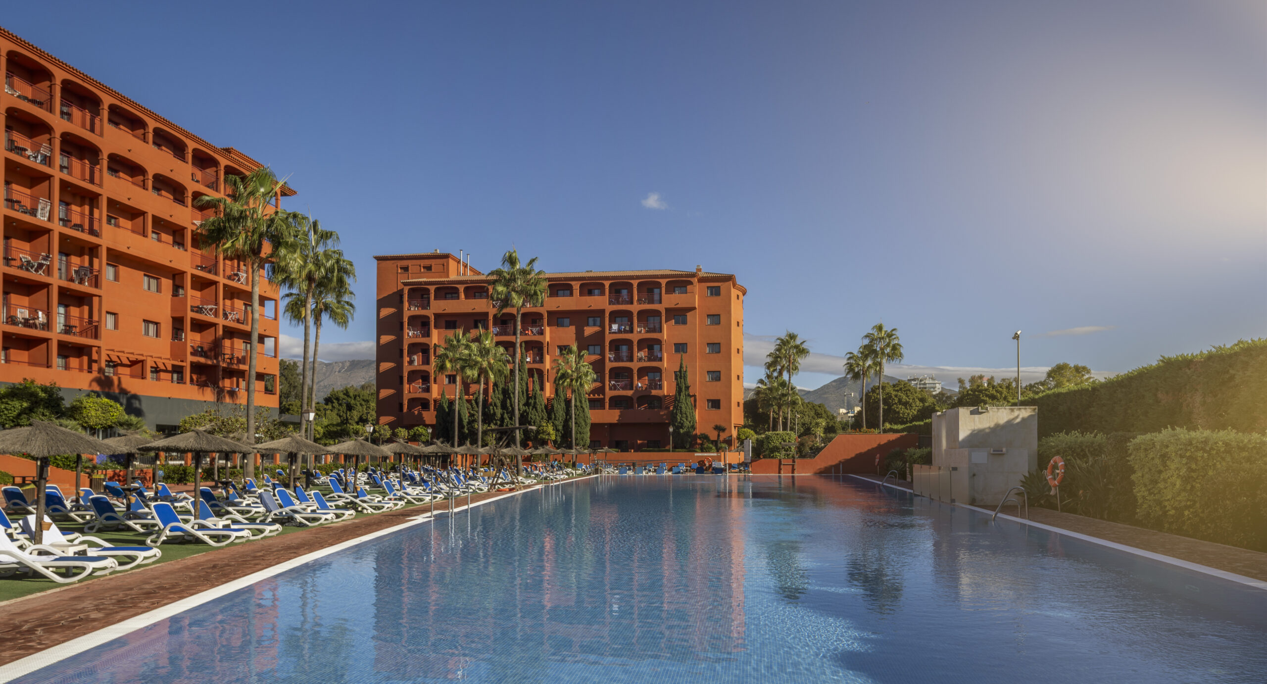 Complejo hotelero con edificios terracota alrededor de una gran piscina, rodeada de hamacas y palmeras bajo cielo despejado.
