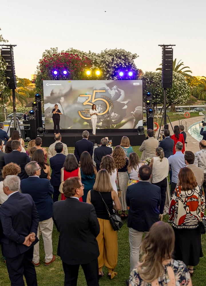 Escenario exterior con pantalla que muestra “35 ANIVERSARIO ILUNION” y una persona presentando, acompañada por otra que interpreta en lengua de signos. Frente al escenario, un público numeroso observa de pie en un jardín con iluminación de colores y árboles alrededor.