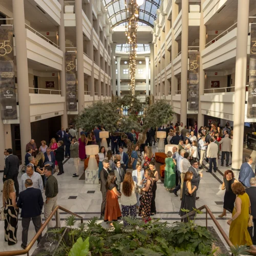 Gran atrio interior luminoso con varias plantas en galerías simétricas y una claraboya superior. En la planta baja, numerosas personas vestidas de forma formal conversan en grupos alrededor de mesas altas y una zona ajardinada central. Del techo cuelga una lámpara alargada, y en las columnas hay banners con el número “35”.