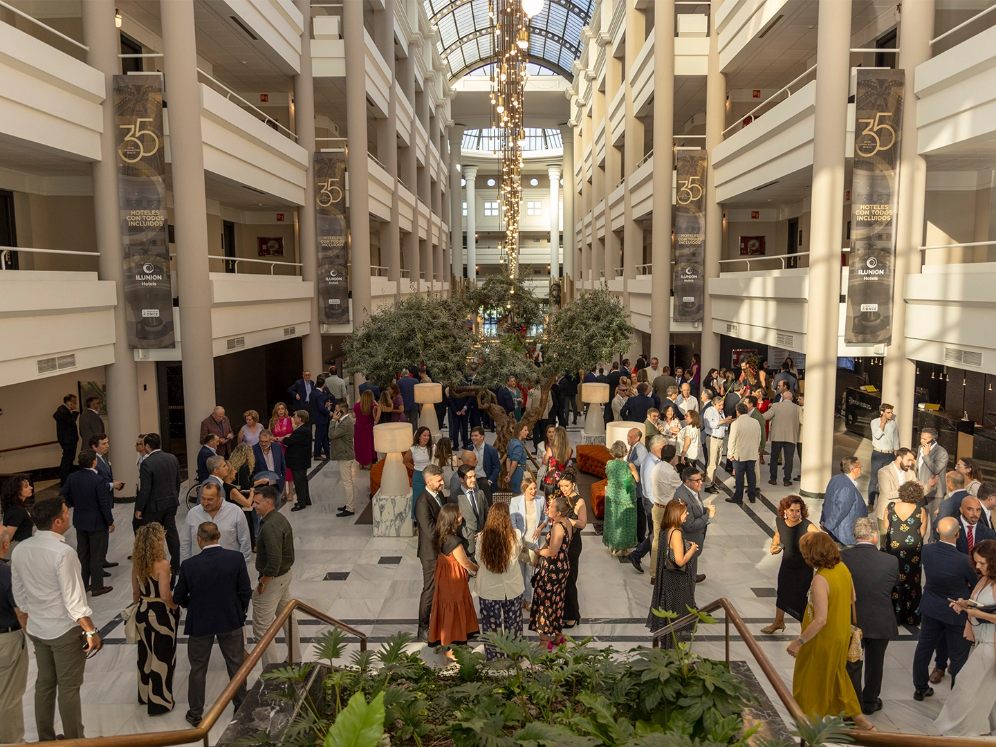 Gran atrio interior luminoso con varias plantas en galerías simétricas y una claraboya superior. En la planta baja, numerosas personas vestidas de forma formal conversan en grupos alrededor de mesas altas y una zona ajardinada central. Del techo cuelga una lámpara alargada, y en las columnas hay banners con el número “35”.