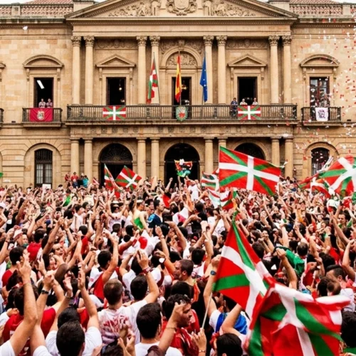 Plaza abarrotada de personas vistas desde atrás y de perfil, con numerosos brazos levantados sosteniendo banderas rojas, verdes y blancas con una cruz blanca y verde. En primer plano, la multitud ocupa casi todo el encuadre, con banderas ondeando a distintas alturas. Al fondo se alza un edificio histórico de piedra con fachada simétrica, columnas y balcones, desde los que también cuelgan varias banderas iguales y otras banderas oficiales. En el balcón central se ven varias personas asomadas observando la escena. La arquitectura es clásica, con arcos en la planta baja y un frontón decorado en la parte superior, y la luz es diurna y uniforme, lo que permite distinguir claramente el conjunto del edificio y la concentración de personas en la plaza.