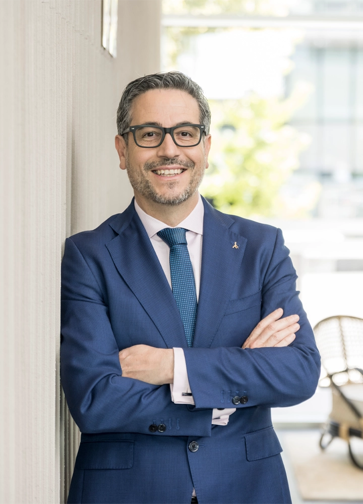Persona adulta apoyada de espaldas a una pared clara en un espacio interior luminoso, con los brazos cruzados a la altura del torso. Viste traje azul oscuro bien ajustado, camisa blanca y corbata azul con textura fina; en la solapa de la chaqueta lleva un pequeño pin metálico. El rostro aparece desenfocado. Al fondo, ligeramente desenfocados, se distinguen grandes ventanales que dejan pasar luz natural, vegetación exterior y parte de una terraza o zona de descanso con una silla y una carretilla. La escena transmite un entorno profesional y cuidado, con iluminación suave y tonos claros.