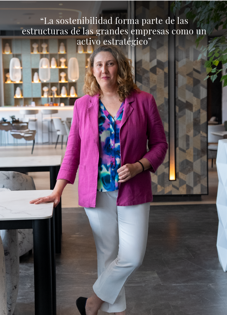 Interior contemporáneo de estilo corporativo con iluminación cálida y diseño cuidado. En primer plano, una persona se sitúa de pie junto a una mesa de superficie clara con patas oscuras, apoyando una mano sobre ella. Lleva chaqueta rosa, blusa estampada en tonos azules, verdes y morados, pantalón claro y calzado oscuro. La postura es relajada, con una pierna ligeramente cruzada por delante de la otra. El fondo muestra una zona tipo cafetería o espacio colaborativo con mesas altas, taburetes y una barra con estanterías iluminadas. Varias lámparas colgantes de formas cilíndricas y ovaladas aportan luz cálida. A la derecha, una pared decorativa con patrón geométrico en tonos madera incorpora una luz vertical integrada. El suelo es oscuro y continuo, contrastando con los muebles claros. En la parte superior aparece el texto: “La sostenibilidad forma parte de las estructuras de las grandes empresas como un activo estratégico”.