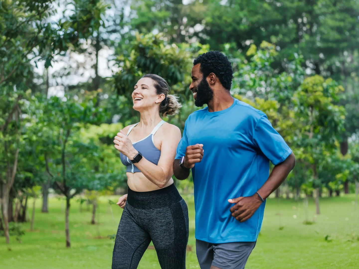 Dos personas corren juntas por un parque verde, avanzando de izquierda a derecha sobre césped bien cuidado, con árboles frondosos y vegetación densa al fondo. Ambas visten ropa deportiva: una lleva un top ajustado y mallas oscuras, y la otra una camiseta amplia azul y pantalones cortos grises; las dos usan relojes deportivos en la muñeca. Los brazos están flexionados en posición de carrera y los cuerpos ligeramente inclinados hacia delante, indicando movimiento. La escena está iluminada por luz natural diurna, con colores vivos y un entorno exterior tranquilo que refuerza una actividad física al aire libre.