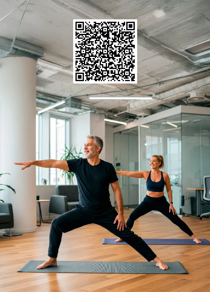 Espacio interior amplio y moderno con suelo de madera clara y paredes blancas, similar a una oficina o sala polivalente. En primer plano, dos personas practican una postura de yoga sobre esterillas grises colocadas paralelas entre sí. La persona situada delante adopta una zancada amplia con una pierna flexionada y la otra estirada hacia atrás, el brazo delantero extendido horizontalmente y el brazo trasero alineado con el cuerpo; la persona situada detrás realiza la misma postura, ligeramente retrasada en el encuadre. Ambas visten ropa deportiva oscura y van descalzas. Al fondo se observan columnas blancas, plantas de interior, paredes acristaladas y mobiliario de oficina, con iluminación natural combinada con luz artificial del techo, creando un entorno ordenado y tranquilo para la actividad física. Con un QR en la parte superior central de la imagen.