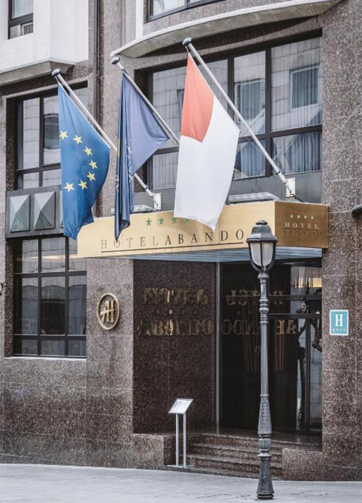 Fachada de un edificio urbano con acabado en piedra donde se sitúa la entrada de un hotel, marcada por una marquesina dorada con el texto “HOTEL ABANDO” y una placa con varias estrellas en el lateral, bajo tres mástiles inclinados que sostienen banderas, incluyendo la bandera de la Unión Europea con estrellas amarillas sobre fondo azul, otra bandera azul y una tercera con bandas claras y una zona superior en tono rojizo; debajo, una puerta acristalada da acceso al interior, acompañada de un pequeño atril informativo en la acera y una farola clásica en primer plano, mientras que las ventanas del edificio reflejan el entorno exterior.