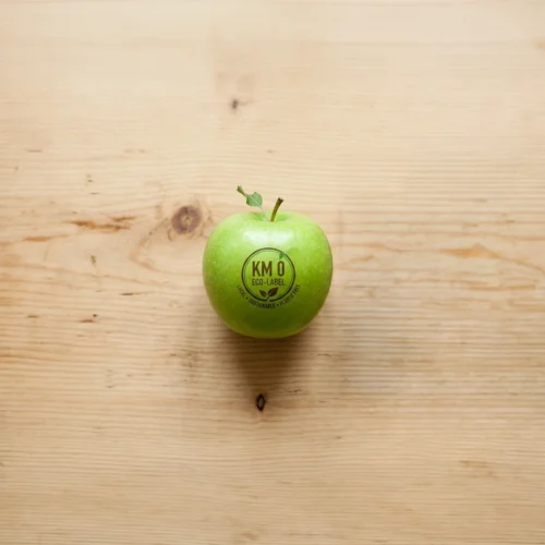 Manzana verde colocada en el centro de una superficie de madera clara, vista desde arriba, con una pequeña hoja en el tallo y un sello circular impreso que dice “KM 0 · ECO · LOCAL”, rodeada de amplio espacio vacío que refuerza una composición limpia y minimalista.
