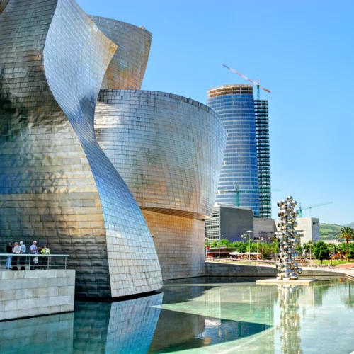 Edificio de arquitectura contemporánea con grandes volúmenes curvos revestidos de paneles metálicos plateados y dorados, situado junto a una lámina de agua que refleja las fachadas; a la izquierda, una pasarela elevada con barandilla donde varias personas caminan o se detienen, y en el centro del estanque una escultura vertical formada por esferas metálicas brillantes apiladas; al fondo aparecen edificios modernos, uno cilíndrico y acristalado con una grúa en la parte superior, zonas verdes con palmeras y un cielo azul despejado que realza los reflejos del metal y el agua.