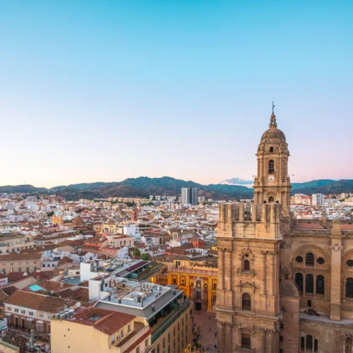 Vista panorámica de una ciudad al atardecer con numerosas edificaciones de baja altura y tejados claros, dominada en primer plano por una gran catedral de piedra con una torre campanario alta y ornamentada; al fondo se extienden colinas suaves bajo un cielo degradado de azul a tonos cálidos, mientras la luz natural ilumina de forma uniforme todo el paisaje urbano.