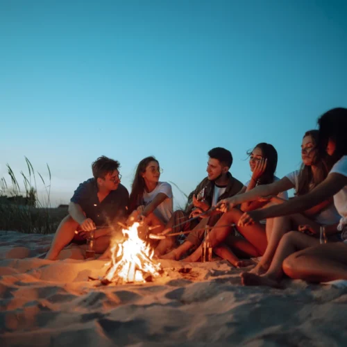 Grupo de varias personas sentadas sobre la arena de una playa al atardecer, rodeando una pequeña hoguera en la que algunas de ellas sostienen palos para acercarlos al fuego; la luz cálida de las llamas ilumina parcialmente los cuerpos y contrasta con el cielo azul tenue, mientras en el entorno se aprecian dunas suaves y vegetación baja, creando una escena tranquila junto al mar.