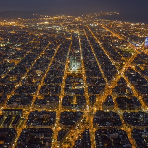 Vista aérea nocturna de una ciudad con cuadrícula muy regular y manzanas octogonales iluminadas por luces cálidas; en el centro destaca una gran basílica de varias torres claramente iluminada y, hacia la derecha, un rascacielos moderno con iluminación de color rompe la uniformidad, mientras la trama urbana se extiende hasta el horizonte donde se intuyen colinas y una masa de agua en la oscuridad.