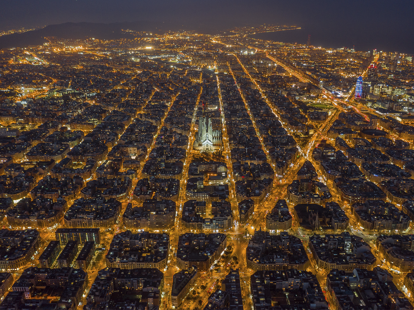 Vista aérea nocturna de una ciudad con cuadrícula muy regular y manzanas octogonales iluminadas por luces cálidas; en el centro destaca una gran basílica de varias torres claramente iluminada y, hacia la derecha, un rascacielos moderno con iluminación de color rompe la uniformidad, mientras la trama urbana se extiende hasta el horizonte donde se intuyen colinas y una masa de agua en la oscuridad.