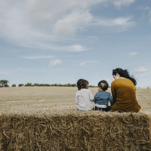 Escena al aire libre en un campo abierto bajo un cielo amplio con nubes dispersas, donde tres personas aparecen sentadas de espaldas sobre un gran fardo de paja en primer plano; las dos figuras más pequeñas se sitúan en el centro con ropa informal de tonos claros y azul, mientras la figura situada a la derecha viste una prenda de color marrón y tiene el cabello recogido, todas apoyadas mirando hacia la extensión del terreno; frente a ellas se extiende una llanura de tonos dorados y tierra seca con una línea de árboles en el horizonte, y la luz suave resalta las texturas de la paja y el paisaje, generando una composición tranquila y equilibrada sin presencia de texto ni elementos adicionales.