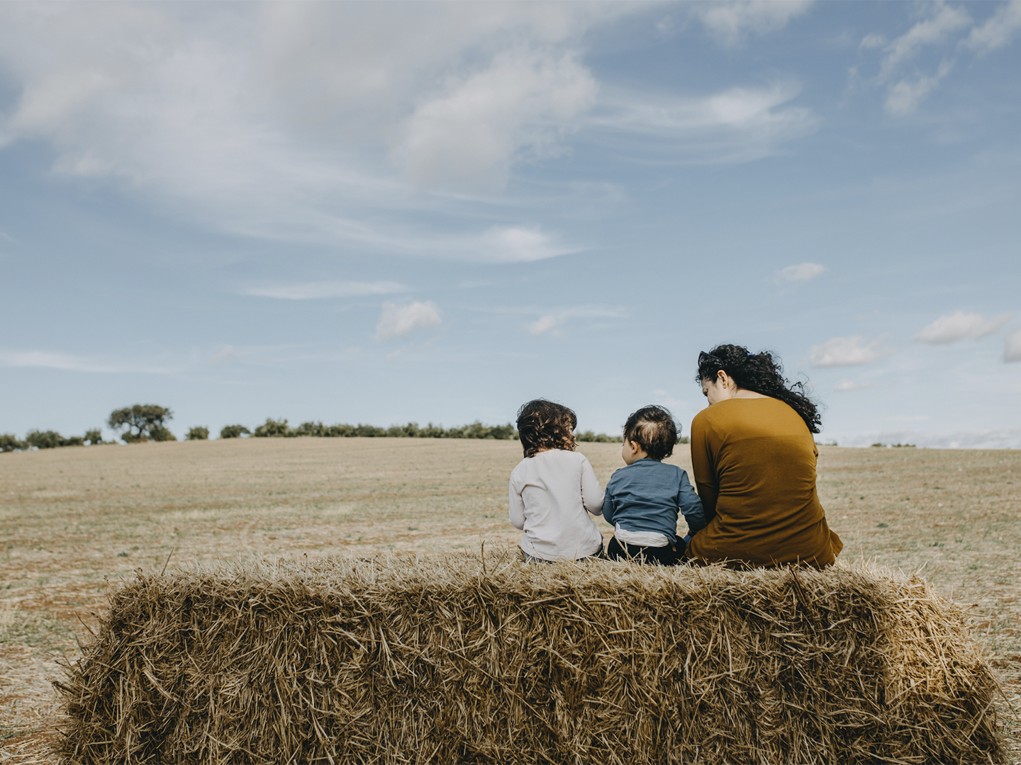 Escena al aire libre en un campo abierto bajo un cielo amplio con nubes dispersas, donde tres personas aparecen sentadas de espaldas sobre un gran fardo de paja en primer plano; las dos figuras más pequeñas se sitúan en el centro con ropa informal de tonos claros y azul, mientras la figura situada a la derecha viste una prenda de color marrón y tiene el cabello recogido, todas apoyadas mirando hacia la extensión del terreno; frente a ellas se extiende una llanura de tonos dorados y tierra seca con una línea de árboles en el horizonte, y la luz suave resalta las texturas de la paja y el paisaje, generando una composición tranquila y equilibrada sin presencia de texto ni elementos adicionales.