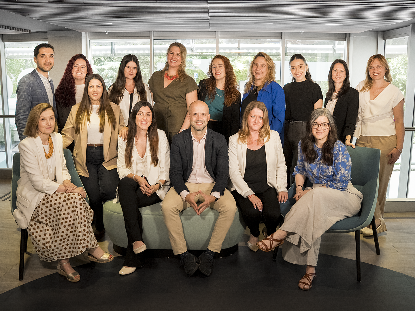 Grupo de quince personas posando en un espacio interior moderno y luminoso, organizadas en dos filas alrededor de asientos tapizados: varias están sentadas en la parte frontal y el resto de pie detrás. Visten ropa formal y semiformal en tonos neutros y algunos colores más vivos. El fondo muestra grandes ventanales que dejan entrar luz natural, con vistas exteriores difuminadas, techo con lamas horizontales y un entorno corporativo ordenado y profesional.