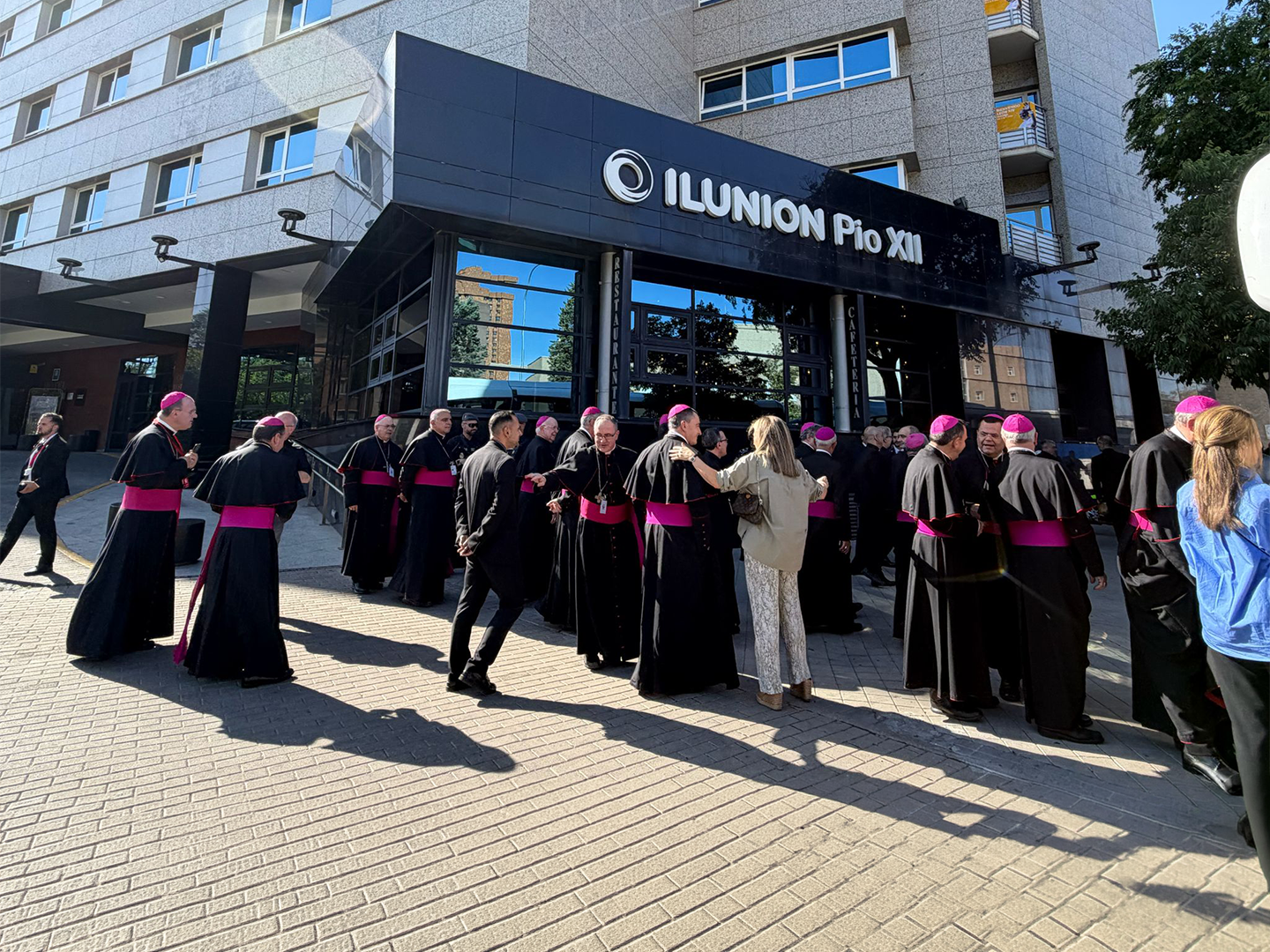 Entrada de un edificio con el rótulo “ILUNION Pío XII” en una marquesina negra; en primer plano, un grupo numeroso de personas con vestimenta religiosa oscura y fajines fucsias se reúne y conversa frente a la puerta, mientras otras personas vestidas de forma informal se mezclan entre el grupo. La fachada es de piedra clara con ventanas y balcones, y la escena está iluminada por luz natural que proyecta sombras sobre el pavimento.