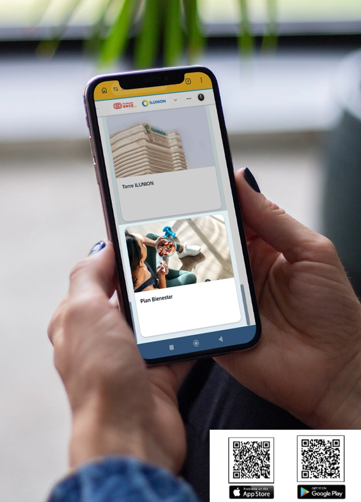 Un teléfono móvil sostenido con ambas manos muestra en la pantalla una aplicación corporativa con dos tarjetas principales: una superior con la imagen de un edificio moderno titulada “Torre ILUNION” y otra inferior con varias personas en una actividad de bienestar bajo el texto “Plan Bienestar”; el fondo aparece desenfocado con luz natural y hojas verdes, y en la parte inferior de la imagen, fuera del teléfono, se ven dos códigos QR acompañados de los iconos de App Store y Google Play para la descarga de la aplicación.
