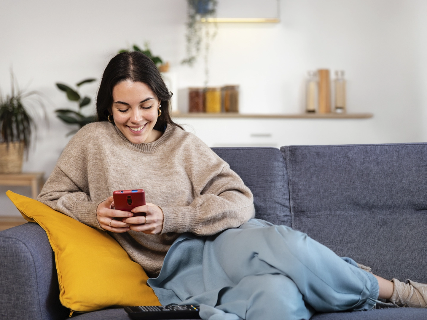 Persona recostada en un sofá de tela gris dentro de un salón doméstico, con las piernas estiradas y apoyadas sobre el asiento, sosteniendo un teléfono móvil rojo con ambas manos; viste ropa cómoda de tonos neutros y claros, con un cojín amarillo colocado a un lado del cuerpo, mientras en primer plano sobre el sofá se aprecia un mando a distancia; el fondo incluye una pared clara con una balda de madera, frascos decorativos y una planta verde, todo iluminado por luz suave y natural que crea un ambiente tranquilo y cotidiano.