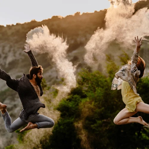 Escena al aire libre en un entorno natural con una ladera rocosa y vegetación al fondo, iluminada por luz cálida de atardecer, donde dos personas aparecen suspendidas en pleno salto con los pies descalzos y las extremidades extendidas; la figura de la izquierda viste camisa de cuadros abierta y pantalones arremangados, con una pierna estirada hacia atrás y otra flexionada, mientras lanza polvo claro que forma una nube densa y alargada sobre la cabeza, y la figura de la derecha lleva una prenda superior ligera y pantalón corto, arquea el cuerpo con ambos brazos elevados y genera otra nube de polvo que se dispersa en el aire, creando trazos fluidos que contrastan con el fondo más oscuro y aportan dinamismo a la escena, sin presencia de texto ni objetos adicionales visibles.