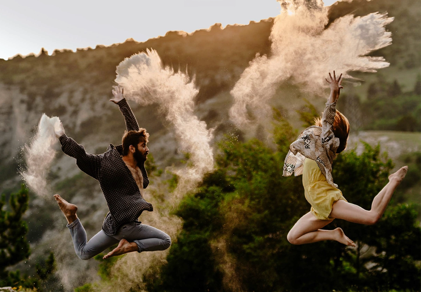 Escena al aire libre en un entorno natural con una ladera rocosa y vegetación al fondo, iluminada por luz cálida de atardecer, donde dos personas aparecen suspendidas en pleno salto con los pies descalzos y las extremidades extendidas; la figura de la izquierda viste camisa de cuadros abierta y pantalones arremangados, con una pierna estirada hacia atrás y otra flexionada, mientras lanza polvo claro que forma una nube densa y alargada sobre la cabeza, y la figura de la derecha lleva una prenda superior ligera y pantalón corto, arquea el cuerpo con ambos brazos elevados y genera otra nube de polvo que se dispersa en el aire, creando trazos fluidos que contrastan con el fondo más oscuro y aportan dinamismo a la escena, sin presencia de texto ni objetos adicionales visibles.