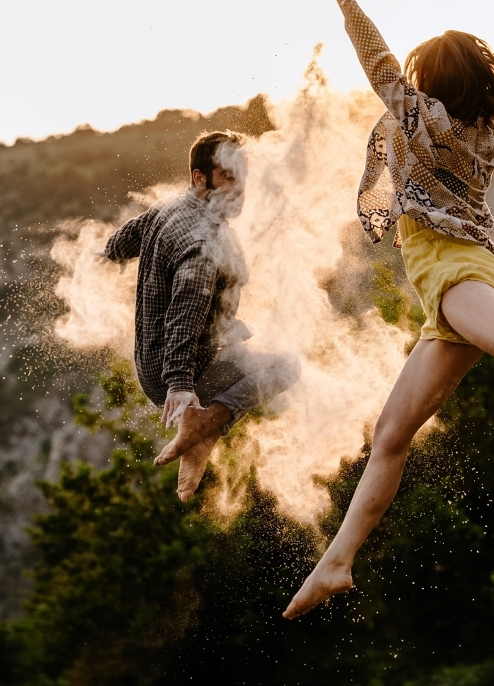 Escena al aire libre en un entorno natural con vegetación y una ladera difusa al fondo, iluminada por luz cálida de atardecer, donde dos personas aparecen suspendidas en pleno salto en primer plano, con los pies descalzos y las piernas flexionadas y extendidas en distintas posiciones, mientras una nube densa de polvo claro o harina estalla alrededor del cuerpo de la persona situada a la izquierda, dispersándose en partículas finas que se iluminan y crean un efecto dinámico y contrastado frente al fondo más oscuro, sin presencia de objetos adicionales ni texto visible.