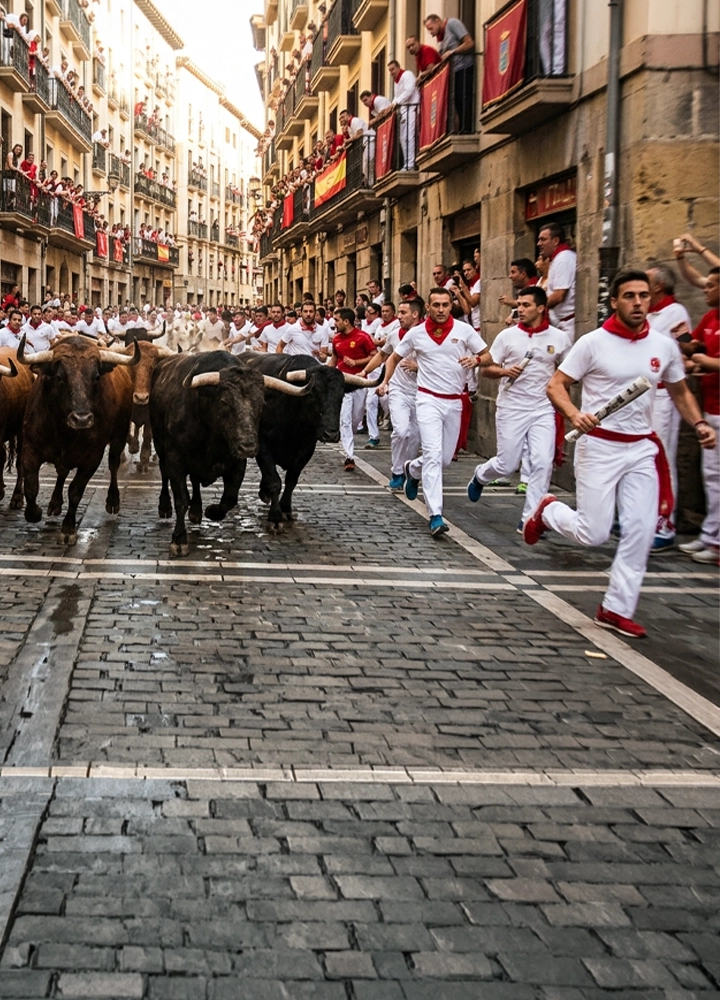 Calle urbana estrecha con suelo de adoquines por la que avanza un grupo de toros de pelaje oscuro y cuernos prominentes, rodeados de numerosas personas vestidas de blanco con pañuelos rojos que corren junto a los animales; en primer plano, varias personas se desplazan a gran velocidad muy cerca de los toros, mientras a ambos lados de la calle los balcones están llenos de espectadores y decorados con telas rojas y banderas, creando un ambiente concurrido y tenso bajo luz de día.