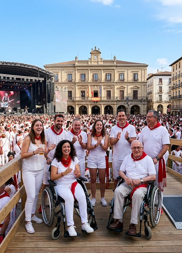Grupo de varias personas reunidas sobre una plataforma de madera en primer plano, algunas de ellas sentadas en sillas de ruedas y otras de pie, todas vestidas de blanco con pañuelos rojos al cuello. Detrás se extiende una gran plaza llena de multitud vestida de forma similar, creando una masa uniforme de blanco y rojo que ocupa todo el espacio hasta el fondo. En el lado izquierdo se ve un escenario elevado con pantallas y una actuación en directo, mientras en el centro del fondo destaca un edificio histórico de fachada simétrica con balcones, un reloj y banderas colgadas en el balcón principal. Los balcones de los edificios laterales también están llenos de personas observando el evento. El suelo de la plaza es de piedra y la iluminación es natural, con un cielo despejado azul. La composición transmite un ambiente festivo y multitudinario en un entorno urbano tradicional.