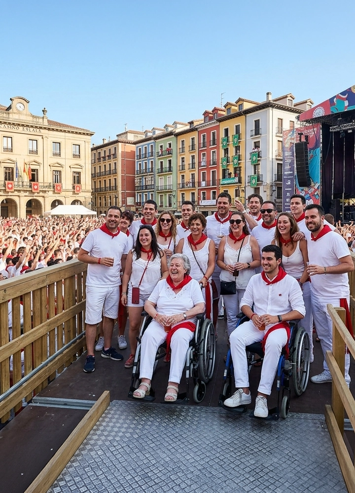 Grupo de personas reunidas sobre una plataforma elevada con barandillas de madera, varias de ellas sentadas en sillas de ruedas y el resto de pie, todas vestidas mayoritariamente de blanco con pañuelos rojos al cuello y algunas sosteniendo bebidas en la mano. Detrás se extiende una plaza completamente llena de gente también vestida de blanco y rojo, creando una gran masa uniforme que ocupa todo el espacio hasta el fondo. A la izquierda se alza un edificio histórico de fachada clara con reloj en la parte superior y banderas colgadas en el balcón principal, mientras a la derecha se ve un escenario de conciertos con estructuras metálicas, pantallas y altavoces. En el fondo aparecen edificios de varios colores —amarillos, verdes, rojos y azules— con balcones llenos de espectadores y decoraciones festivas. El cielo está despejado y la luz natural ilumina la escena, resaltando el ambiente multitudinario y festivo en un entorno urbano tradicional.