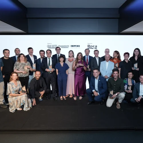 Grupo amplio de unas veinte personas posando sobre un escenario negro frente a un fondo blanco con logotipos “GRAN HOTEL”, “Equipamiento hostelero”, “CLUBT” y “Roca”. Varias personas sostienen trofeos; están organizadas en dos filas, una de pie y otra en cuclillas o sentada, con vestimenta formal de trajes y vestidos de distintos colores.