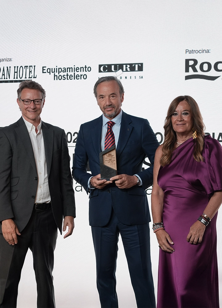 Escenario de entrega de premios con fondo blanco cubierto de logotipos de patrocinadores y organizadores, entre ellos “GRAN HOTEL”, “Equipamiento hostelero”, “CLUBT” y “Roca”. En primer plano, tres personas posan de pie alineadas horizontalmente. La persona situada en el centro viste traje azul oscuro con corbata a rayas rojas y azules y sostiene un trofeo de base oscura con placa metálica. A la izquierda, otra persona lleva traje oscuro con camisa clara sin corbata. A la derecha, una tercera persona viste un vestido brillante en tono morado con pulseras en la muñeca. Iluminación uniforme y fondo limpio típico de photocall corporativo.