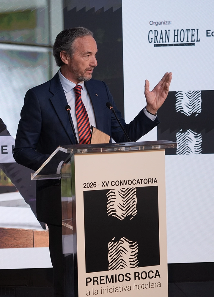 Escenario de conferencias con atril transparente y dos micrófonos inclinados hacia la persona oradora. En el frontal del atril se lee “2026 · XV CONVOCATORIA” y debajo “PREMIOS ROCA a la iniciativa hotelera”, acompañado de un diseño gráfico en negro. La persona detrás del atril viste traje azul y corbata a rayas rojas y sostiene un objeto tipo galardón en la mano izquierda mientras eleva la mano derecha en gesto de intervención. En el fondo aparecen paneles con logotipos, incluido “GRAN HOTEL”, y zonas desenfocadas con colores verde y gris.