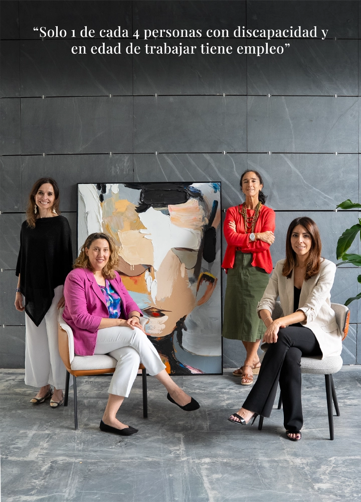 Grupo de cuatro personas posando en un espacio interior con suelo de hormigón y pared de grandes paneles grises; dos están sentadas en sillas tapizadas en primer plano y otras dos permanecen de pie detrás. En el centro, apoyado en el suelo, hay un cuadro grande de estilo abstracto con un rostro formado por pinceladas en tonos beige, negro, blanco y naranja. La persona sentada a la izquierda viste chaqueta rosa, pantalón claro y zapatos oscuros, mientras la persona sentada a la derecha lleva chaqueta clara y pantalón oscuro; ambas adoptan postura relajada en sus sillas. Detrás, una persona viste un conjunto oscuro y otra una prenda roja con falda verde, ambas de pie y alineadas con la composición. A la derecha del cuadro aparece una planta de interior con hojas verdes. En la parte superior, sobre la pared gris, se lee en texto blanco: “Solo 1 de cada 4 personas con discapacidad y en edad de trabajar tiene empleo”. La iluminación es uniforme y suave, resaltando los colores de la ropa y del cuadro sobre el fondo neutro.