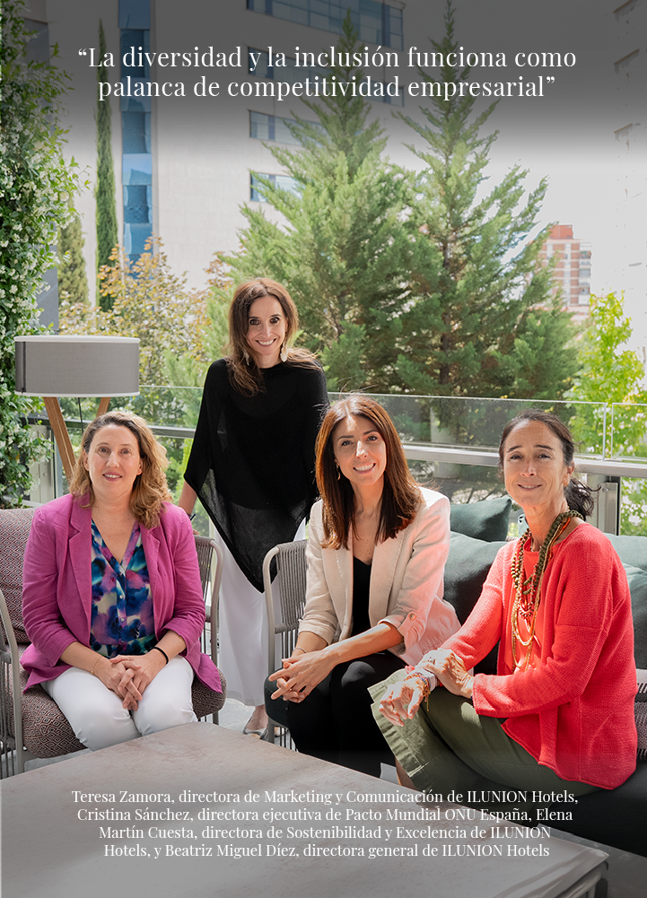 Terraza exterior luminosa con barandilla de cristal y abundante vegetación al fondo, incluyendo árboles de hoja perenne y edificios cercanos difuminados. Cuatro personas se agrupan alrededor de una mesa baja: dos están sentadas en sillones de exterior en primer plano y dos permanecen de pie detrás. La persona sentada a la izquierda viste chaqueta rosa, blusa multicolor y pantalón claro; la persona sentada a la derecha lleva chaqueta clara y pantalón oscuro. Detrás, una figura con prenda superior negra y pantalón claro se sitúa a la izquierda, y otra con chaqueta de tono rojizo y falda verde aparece a la derecha. Sobre la mesa hay una superficie clara sin elementos destacados visibles. En la parte superior se lee: “La diversidad y la inclusión funciona como palanca de competitividad empresarial”. En la parte inferior aparece un texto con varios nombres y cargos vinculados a ILUNION Hotels y Pacto Mundial ONU España.
