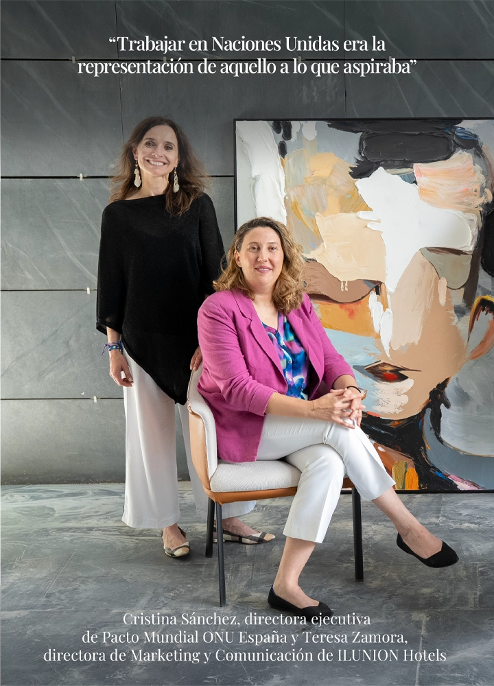 Dos personas posan en un espacio interior con suelo de piedra y pared gris; una está sentada en una silla con patas negras y asiento claro, vestida con chaqueta rosa, pantalón claro y calzado oscuro, con las manos entrelazadas sobre las piernas, mientras otra persona permanece de pie junto al respaldo, vestida con prenda superior negra y pantalón claro. Detrás hay un gran cuadro con un rostro abstracto en tonos beige, negro y naranja. En la parte superior se lee: “Trabajar en Naciones Unidas era la representación de aquello a lo que aspiraba”. En la parte inferior aparece el texto: “Cristina Sánchez, directora ejecutiva de Pacto Mundial ONU España y Teresa Zamora, directora de Marketing y Comunicación de ILUNION Hotels”.