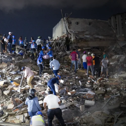 Escena nocturna en una zona urbana con varios edificios residenciales alrededor, donde un gran bloque de hormigón aparece parcialmente colapsado en el centro, convertido en un montón de escombros de ladrillos, vigas y fragmentos de construcción. Sobre los restos, un grupo numeroso de personas se distribuye a distintas alturas, algunas con cascos, chalecos reflectantes o linternas, mientras otras visten ropa casual; muchas están inclinadas, retirando piezas o buscando entre los restos con las manos y herramientas. En primer plano, varias personas se agachan para apartar cascotes y fragmentos de cerámica, mientras otras observan o se desplazan por el terreno irregular. La iluminación artificial procedente de focos y linternas crea zonas muy iluminadas contrastando con sombras profundas, destacando el polvo, los materiales rotos y la textura caótica del terreno. Al fondo, las fachadas de los edificios cercanos permanecen en pie, con algunas ventanas iluminadas, lo que acentúa el contraste entre la estructura intacta y la zona devastada.