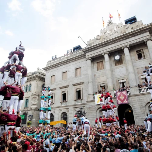 Plaza llena de gente frente a un edificio histórico con columnas y reloj. Varias torres humanas se elevan sobre la multitud, formadas por grupos con camisas de distintos colores, mientras el público sostiene las bases con los brazos en alto.