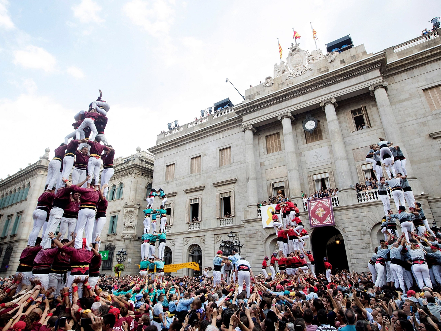 Plaza llena de gente frente a un edificio histórico con columnas y reloj. Varias torres humanas se elevan sobre la multitud, formadas por grupos con camisas de distintos colores, mientras el público sostiene las bases con los brazos en alto.