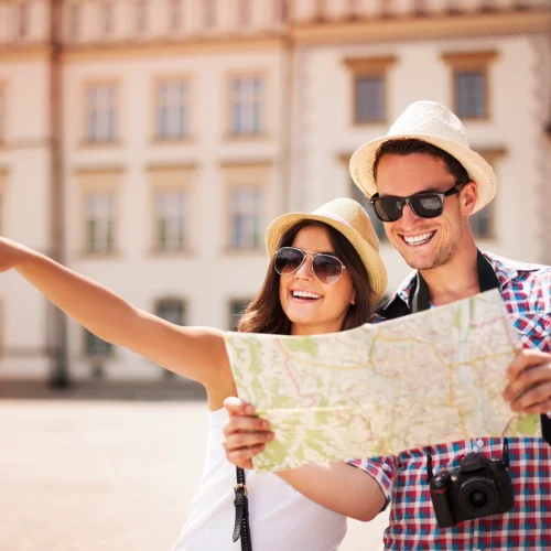 Dos personas en una plaza sostienen un mapa desplegado mientras una extiende el brazo señalando una dirección. Ambas llevan sombreros claros, gafas de sol y ropa de verano. Una cámara cuelga del cuello de una de ellas. Al fondo se observa un edificio histórico con ventanas uniformes, iluminado por luz natural.