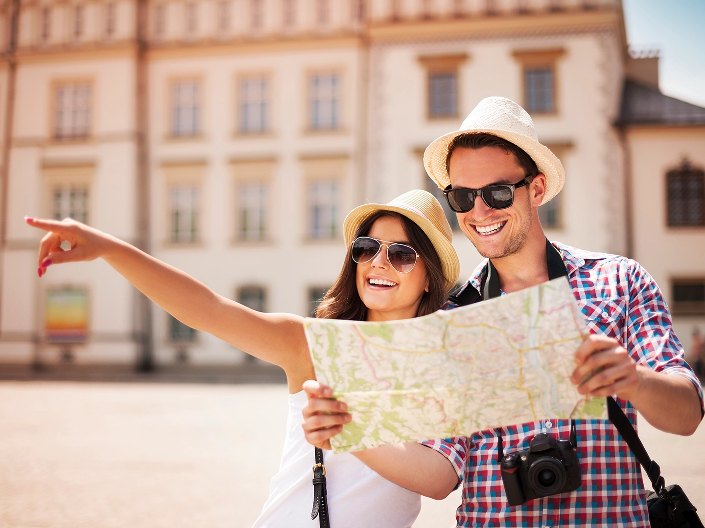 Dos personas en una plaza sostienen un mapa desplegado mientras una extiende el brazo señalando una dirección. Ambas llevan sombreros claros, gafas de sol y ropa de verano. Una cámara cuelga del cuello de una de ellas. Al fondo se observa un edificio histórico con ventanas uniformes, iluminado por luz natural.