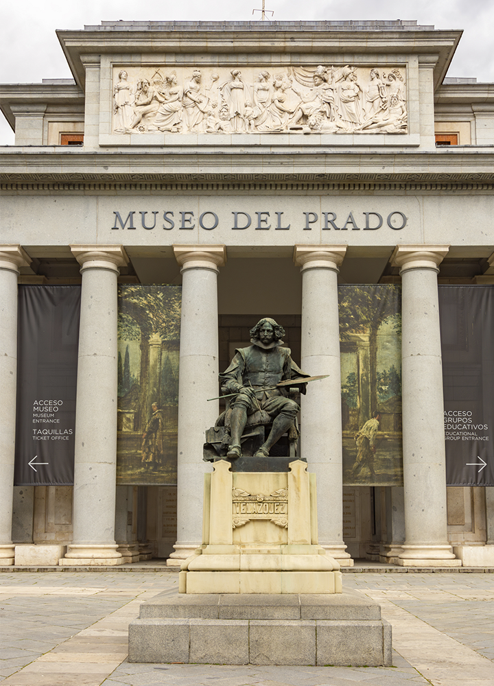Fachada principal del Museo del Prado construida en piedra clara con un pórtico de columnas clásicas que sostienen un frontón decorado con relieves escultóricos de figuras humanas en movimiento, en cuyo centro se lee en letras grandes “MUSEO DEL PRADO”; frente a la entrada se alza una estatua de un personaje sentado sobre un pedestal, con un objeto alargado apoyado sobre las piernas, situada sobre una escalinata de piedra, mientras a ambos lados de la puerta cuelgan grandes lonas verticales con imágenes y texto relacionado con el acceso al museo, todo bajo una luz suave que resalta la textura de la arquitectura y el tono beige del edificio.
